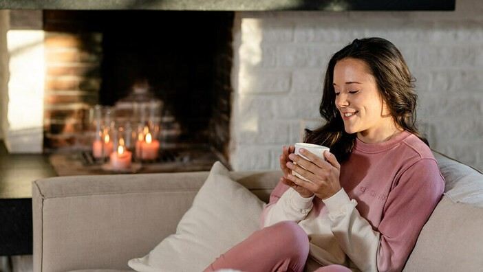 Frau im Pulli sitzt auf dem Sofa mit einer Tasse in der Hand und lächelt