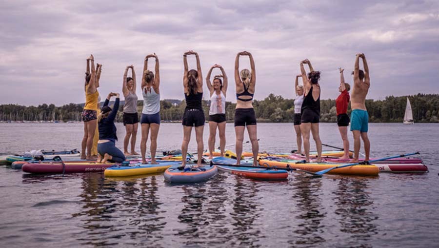 Yoga Sound Sea Festival. Eine Gruppe Frauen steht auf Paddleboards im See und macht Yoga.